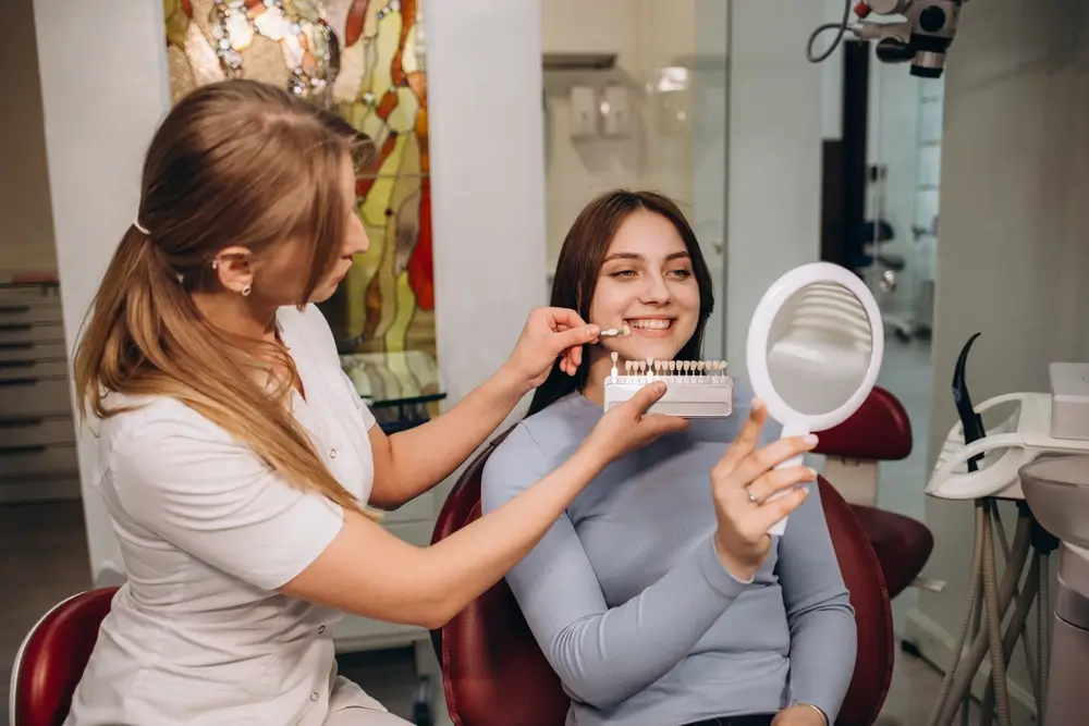 A dentist using a VITA Teeth Color Chart to match the tooth shade of a smiling female patient holding a hand mirror in a bright dental clinic.